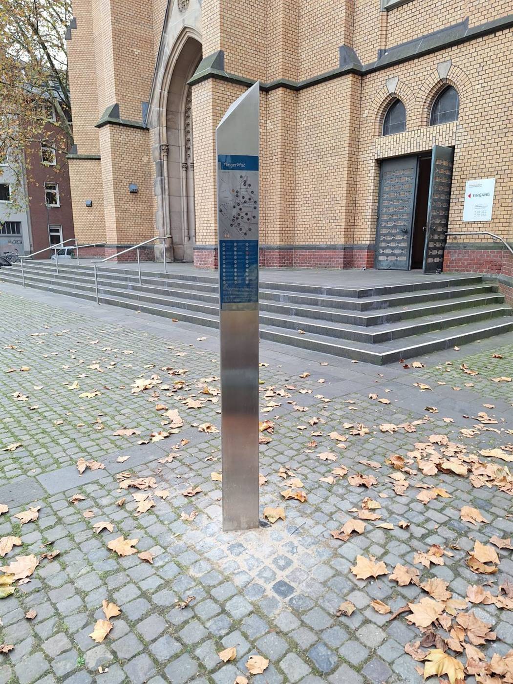 Eine Stele des FlingerPfads mit Übersichtskarte steht auf dem herbstlichen Vorplatz einer Backsteinkirche.